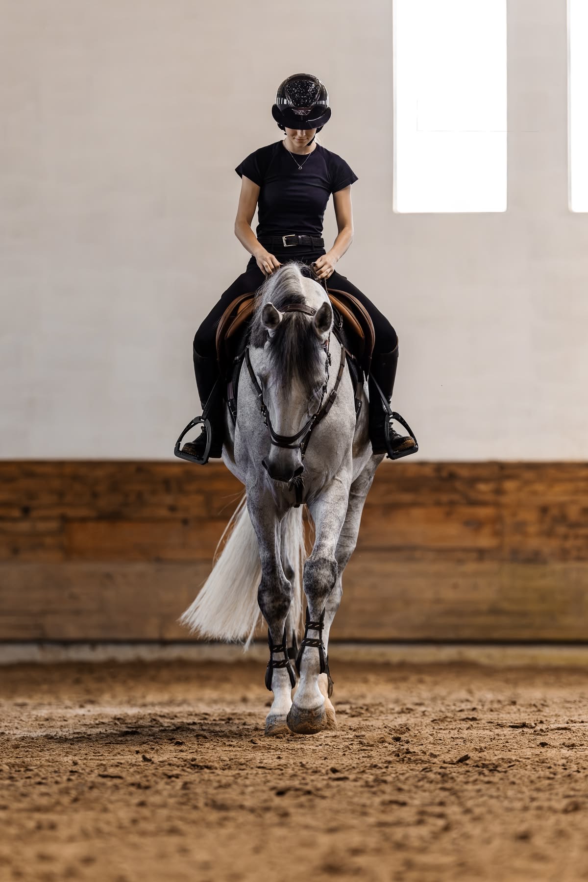 Rider dressed in black on a grey horse, walking toward the camera in an indoor arena
